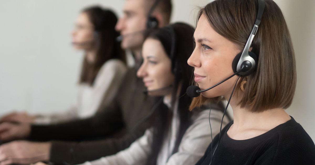 Side view of young coworkers in casual clothes sitting in light workplace with headphones with microphone while surfing on laptop